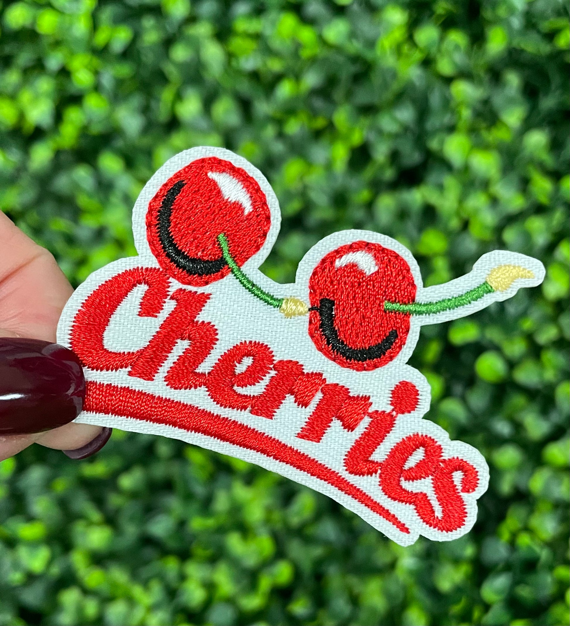An iron-on patch with an embroidered design of two cherries and the word 'Cherries' below them in red and green colors, placed on a bed of green leaves.