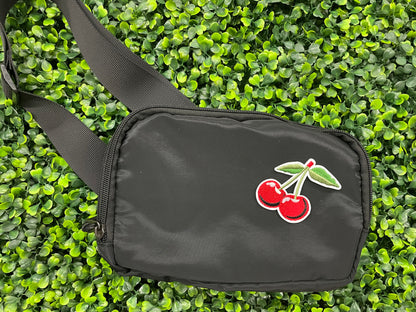 A patch with two red cherries and a green leaf embroidered on it, placed on a black shoulder bag
