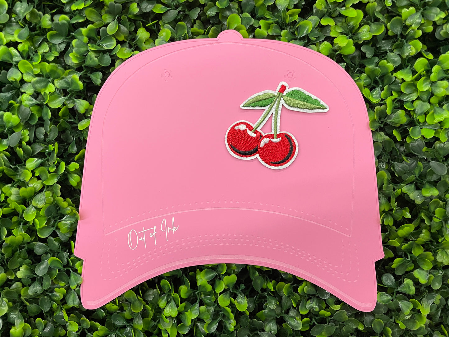 A patch with two red cherries and a green leaf embroidered on it, placed on a pink hat