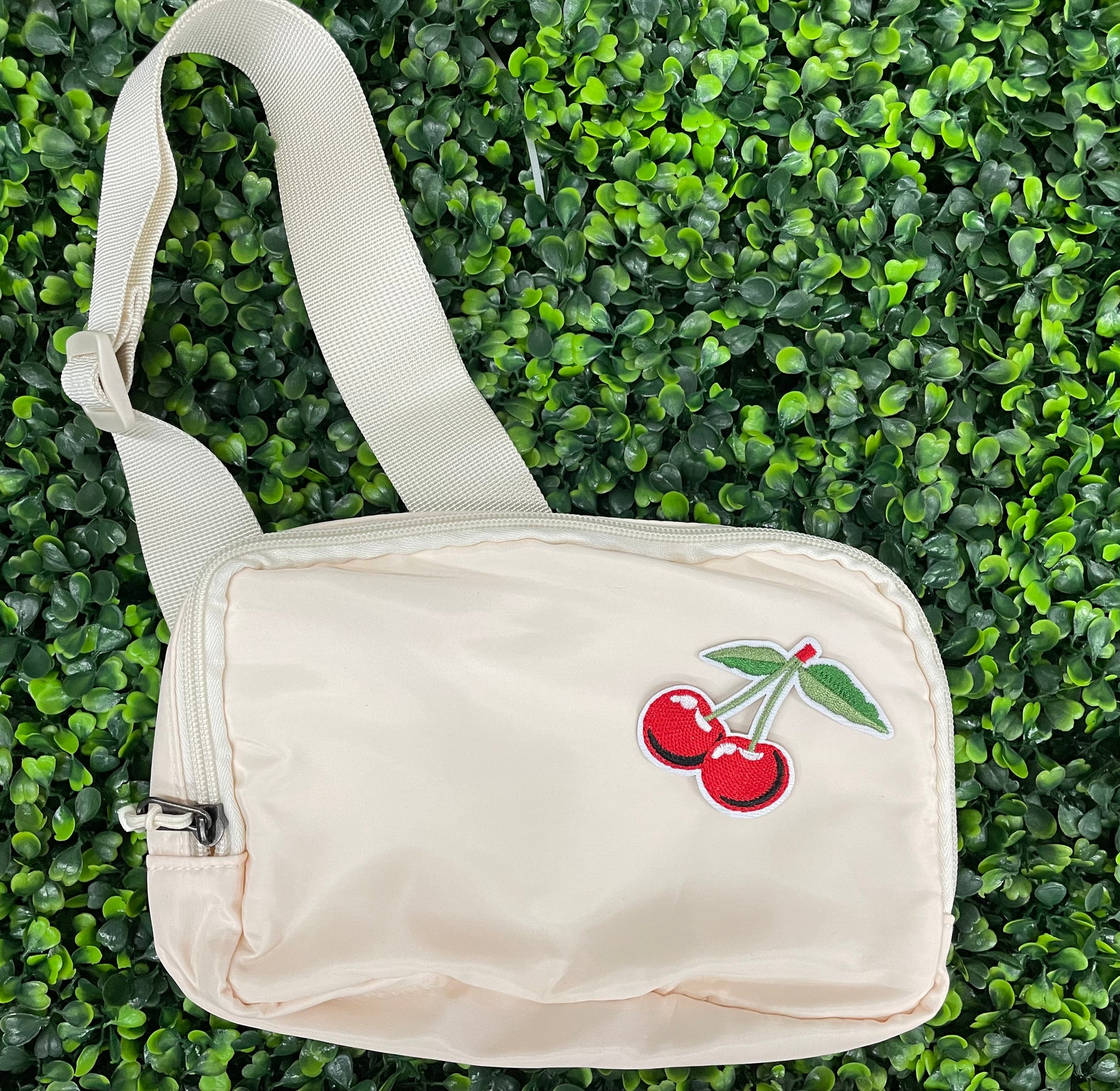 A patch with two red cherries and a green leaf embroidered on it, placed on a white shoulder bag