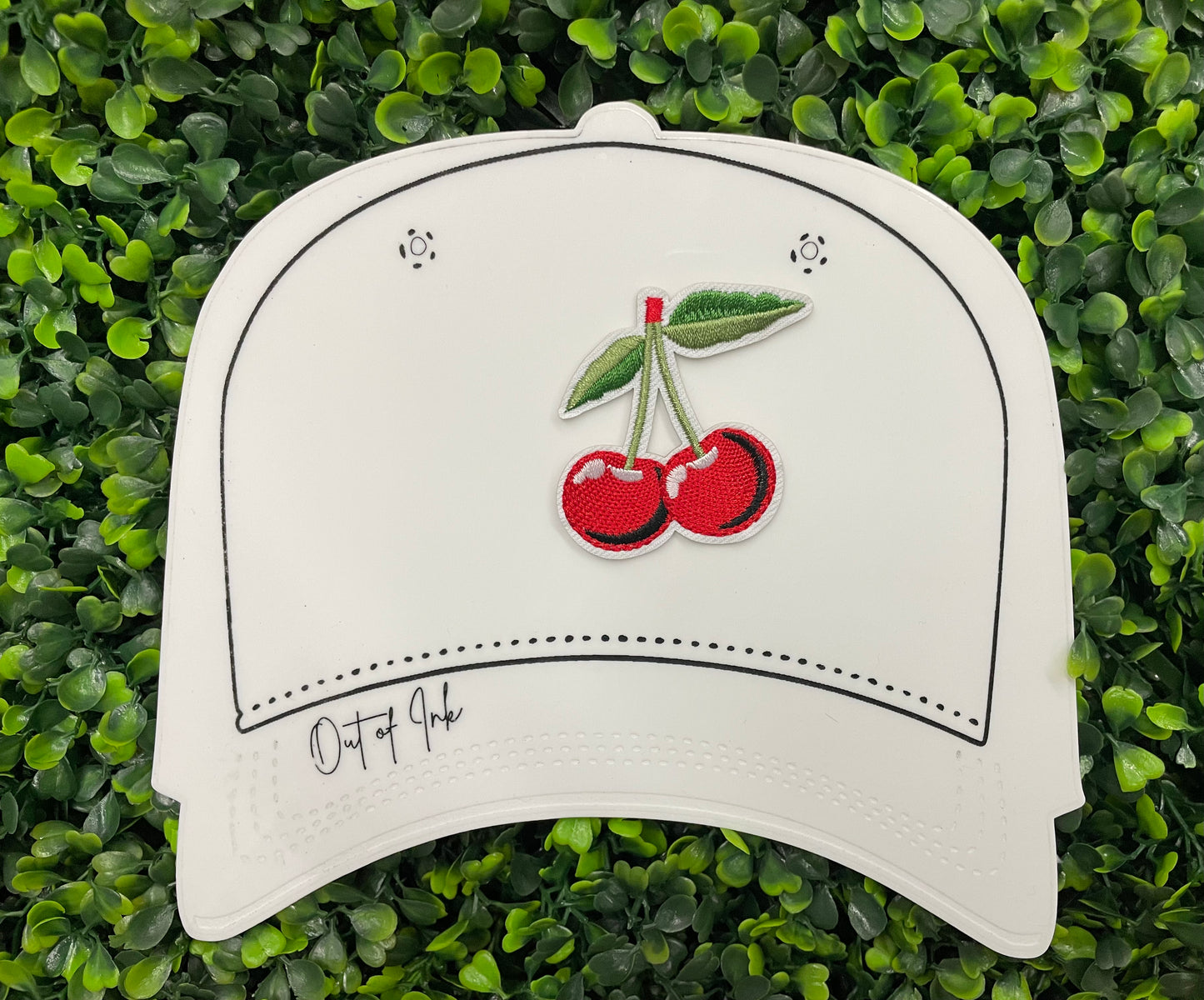 A patch with two red cherries and a green leaf embroidered on it, placed on a white hat graphic