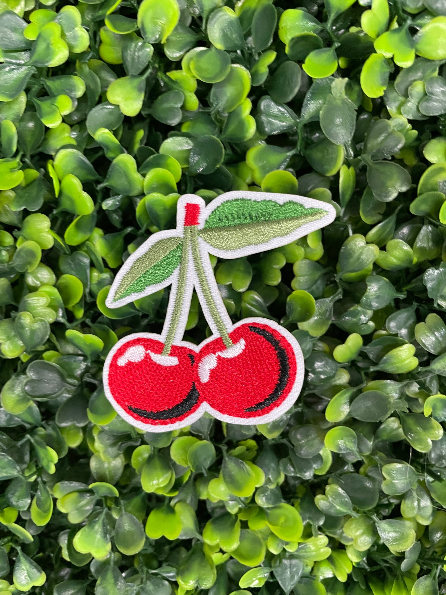 A patch with two red cherries and a green leaf embroidered on it, placed on a greenery background.