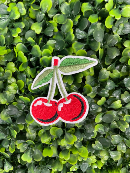 A patch with two red cherries and a green leaf embroidered on it, placed on a greenery background.