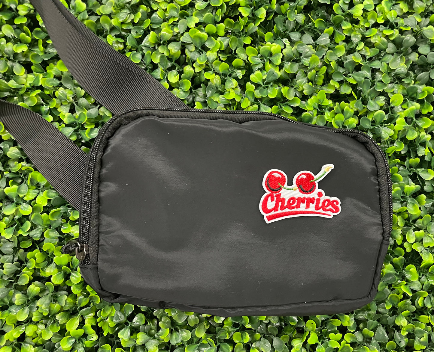 An iron-on patch with an embroidered design of two cherries and the word 'Cherries' below them in red and green colors, placed on a black shoulder bag