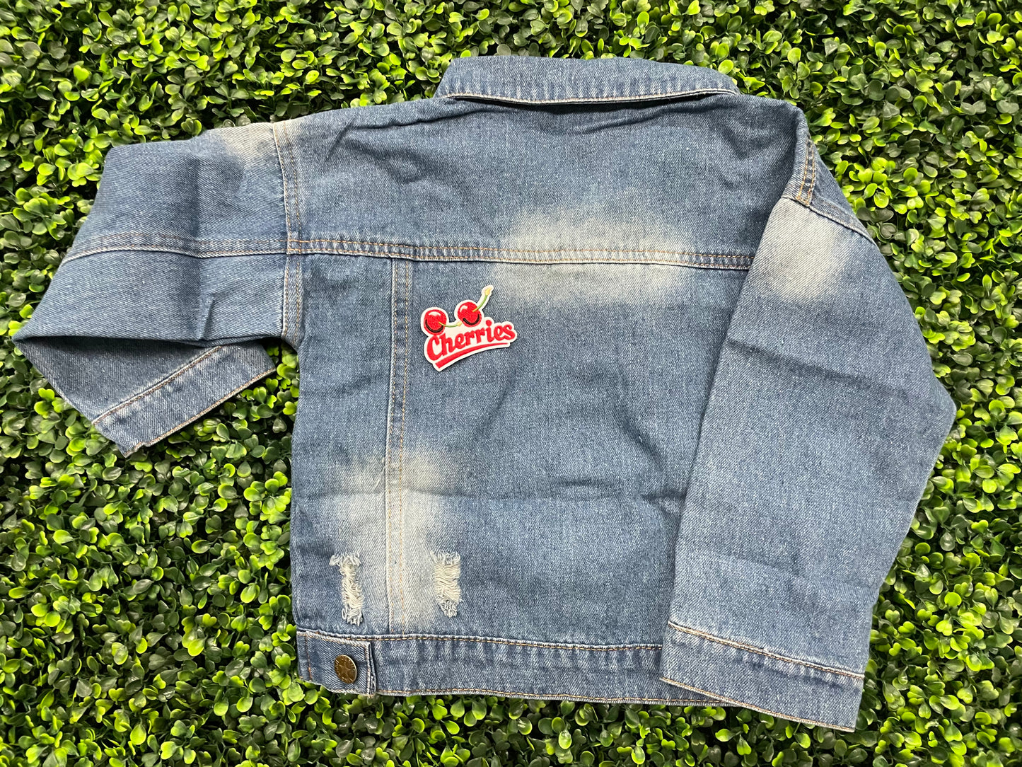 An iron-on patch with an embroidered design of two cherries and the word 'Cherries' below them in red and green colors, placed on a jean jacket