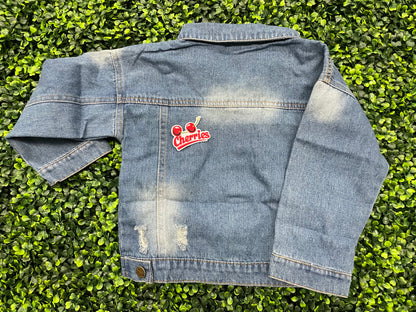 An iron-on patch with an embroidered design of two cherries and the word 'Cherries' below them in red and green colors, placed on a jean jacket