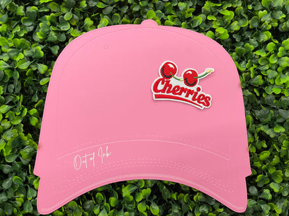 An iron-on patch with an embroidered design of two cherries and the word 'Cherries' below them in red and green colors, placed on a pink hat graphic