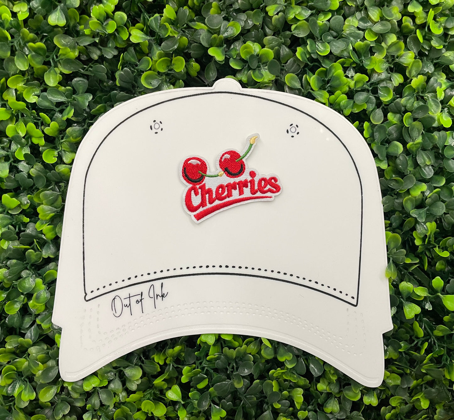 An iron-on patch with an embroidered design of two cherries and the word 'Cherries' below them in red and green colors, placed on a white hat graphic